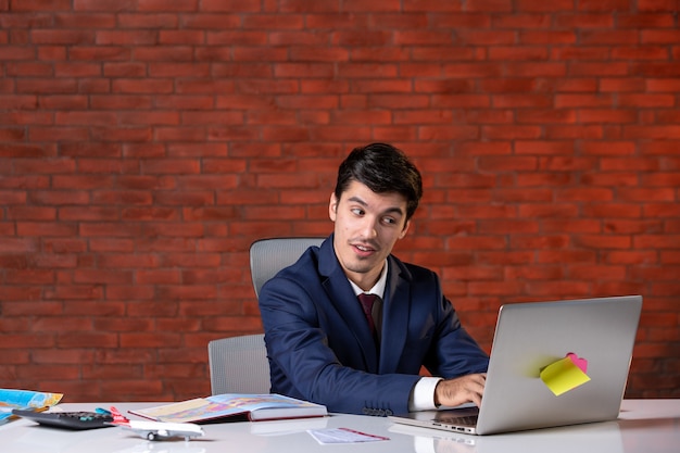 front view of travel agent sitting behind his working place in suit and using laptop corporate occupation service tourism overseas global agency assistant project