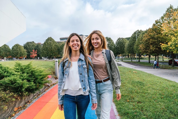 Front view of two cheerful women walking on a path painted with the colors of the rainbow while looking at camera.