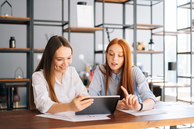 Front view of two happy young business women working using digital tablet during meeting at desk with job documents at office Business partners using touchscreen computer for project discussion