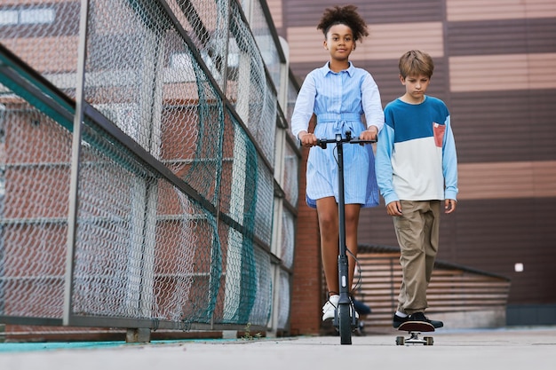 Front view of two intercultural kids riding scooter and skateboard