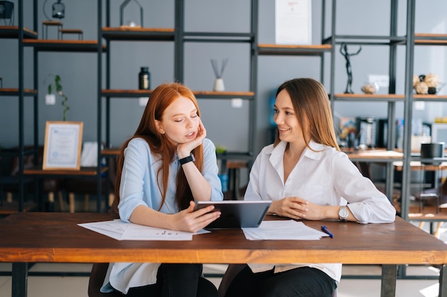 Front view of two relaxed young business women working using digital tablet at meeting desk with job documents at office Business female colleagues using touchscreen computer for project discussion