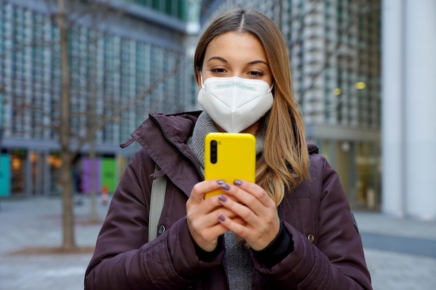 Front view of winter woman with FFP2 KN95 protective mask chatting with smartphone in city street