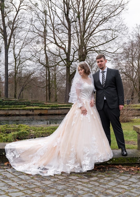 Front view of a young bride dressed in a long very beautiful wedding dress looking away a young groom is standing nearby looking at the camera while walking