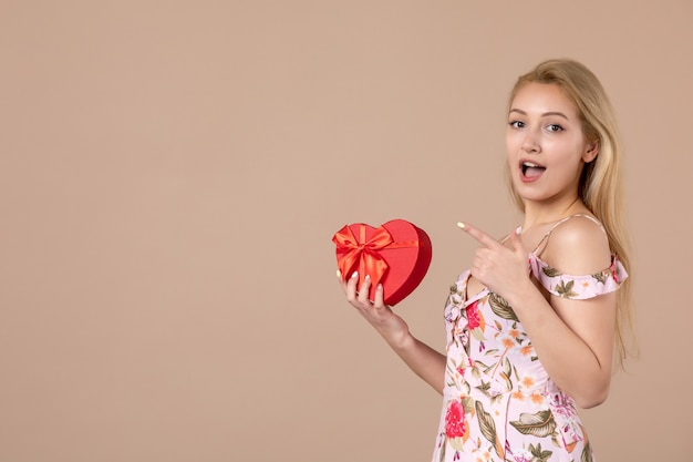 Photo front view of young female posing with red heart shaped present on brown wall