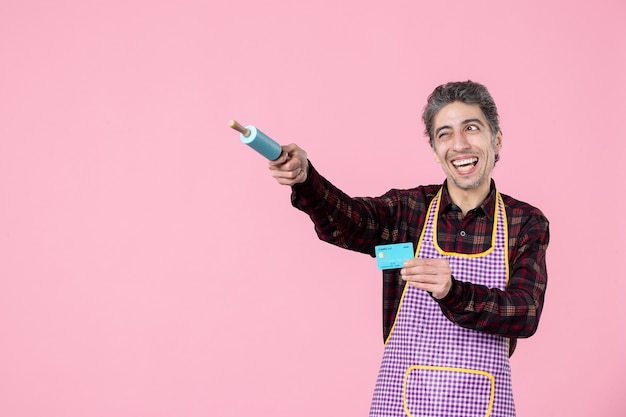 front view young male in cape holding bank card and pointing with little rolling pin on pink background job worker profession horizontal uniform husband