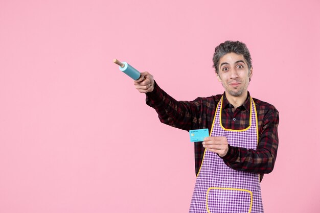 front view young male in cape holding bank card and pointing with little rolling pin on pink background job worker profession horizontal uniform money husband color