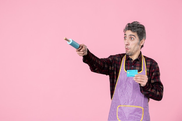 front view young male in cape holding bank card and pointing with little rolling pin on pink background job worker profession uniform money husband