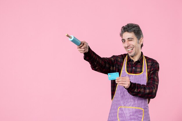 front view young male in cape holding bank card and pointing with little rolling pin on pink background worker profession horizontal uniform money husband