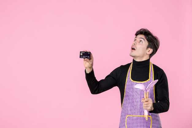 Front view young male in cape holding bank card and spoons on pink background job cooking worker color money horizontal cuisine profession chief