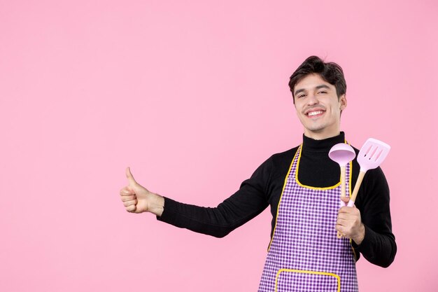 Front view young male in cape holding pink spoons on pink background dough profession cuisine uniform color horizontal chief cooking worker