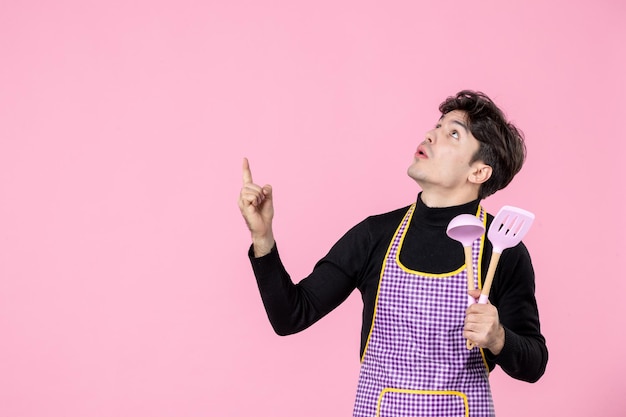 Front view young male in cape holding pink spoons on pink background dough profession uniform color horizontal chief cooking job worker