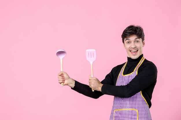 Front view young male in cape holding spoons on pink background job dough profession cuisine uniform cooking worker chief color horizontal