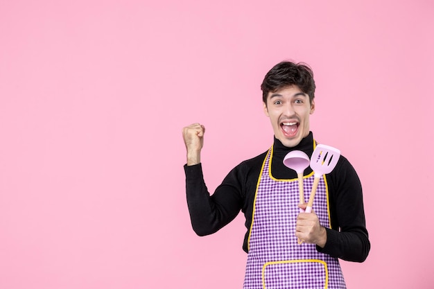 Front view young male in cape holding spoons and rejoicing on pink background dough profession cuisine uniform horizontal chief cooking job color worker