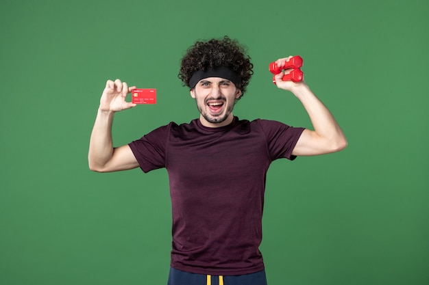 front view young male holding dumbbells and bank card on green background athlete workout sport gym model health shopping colors