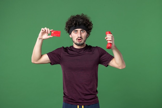 front view young male holding dumbbells and bank card on green background workout sport gym model health athlete shopping color