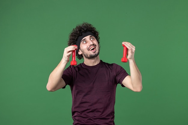 front view young male holding red dumbbells on green background color sport gym emotion health run model athlete lifestyle