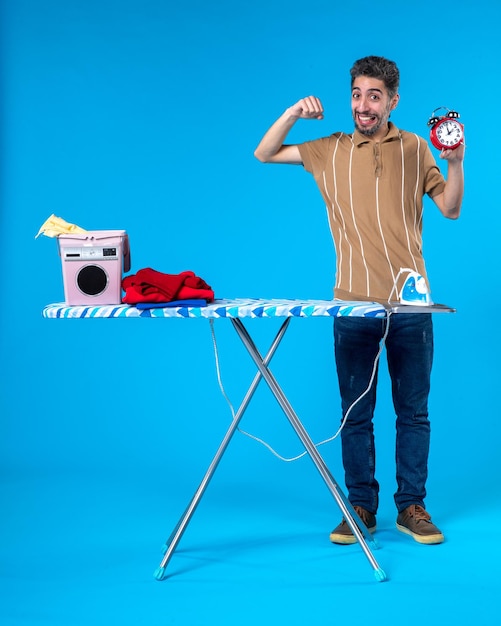 front view young male behind ironing board holding clocks on blue background housework washing machine clean iron time color clothes