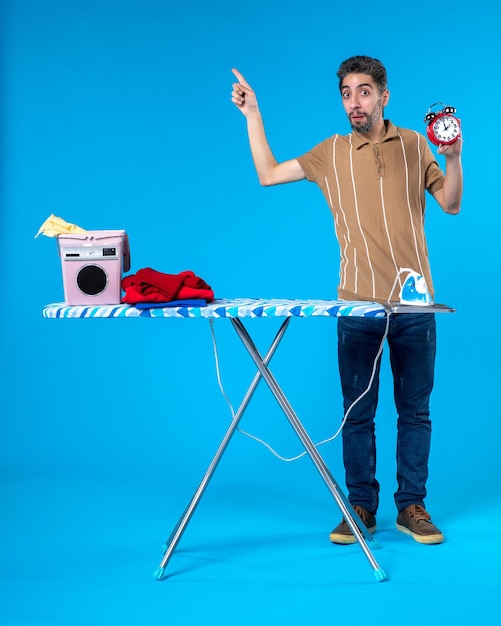 front view young male behind ironing board holding clocks on blue background housework washing machine clean laundry iron time colors