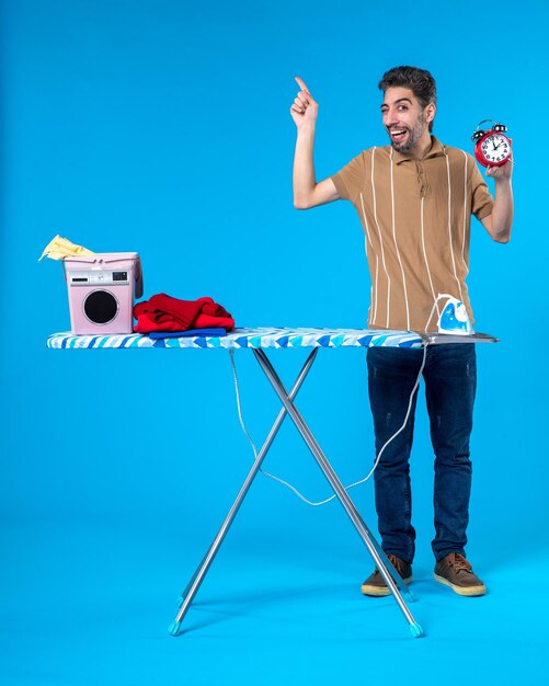 front view young male behind ironing board holding clocks on blue background time housework washing machine clean laundry color