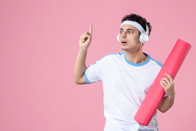 Front view young male in sport clothes with yoga mat and headphones on pink wall