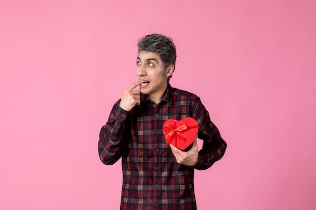 Front view young man holding red heart shaped present on pink wall