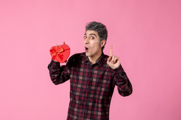 Front view young man holding red heart shaped present on pink wall