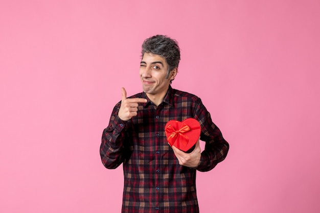 Front view young man holding red heart shaped present on pink wall