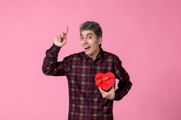 Front view young man holding red heart shaped present on pink wall