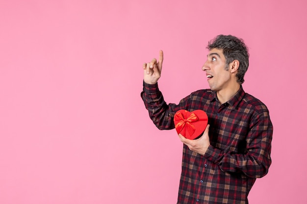 Front view young man holding red heart shaped present on pink wall