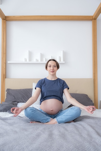 Front view of young pregnant woman sitting in bed in lotus position practicing meditation