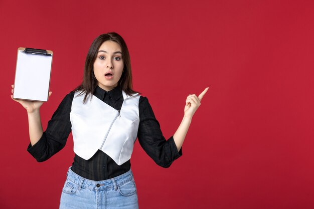 front view young pretty waitress in uniform holding little notepad for orders on red background food color work beauty worker dinner evening job woman