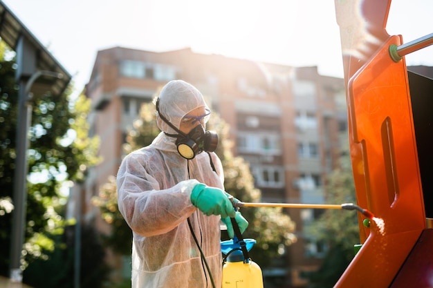 Frontline male worker with face mask and protective workwear using pressure sprayer to disinfect children's playground. Virus pandemic protection and prevention concept.