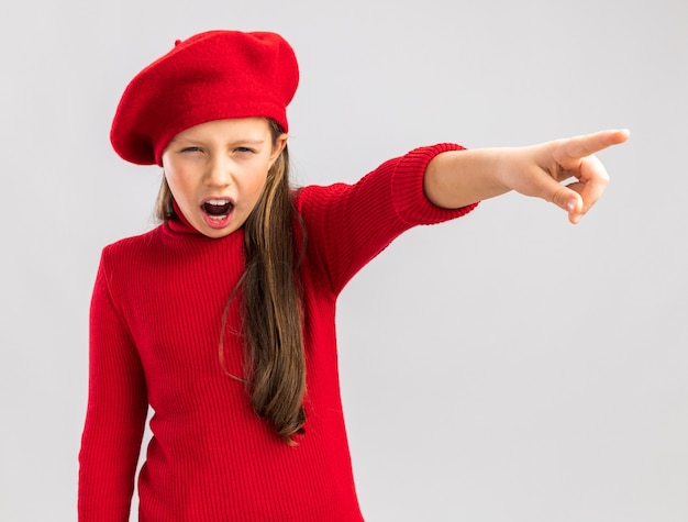 Frowning little blonde girl wearing red beret looking at camera pointing at side isolated on white wall