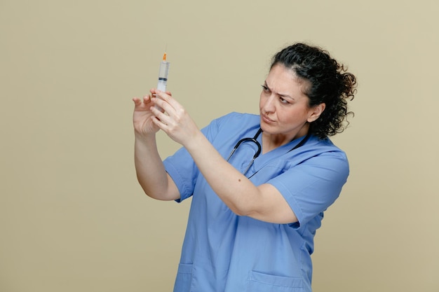 Frowning middleaged female doctor wearing uniform and stethoscope around neck standing in profile view removing air bubbles from syringe isolated on olive background