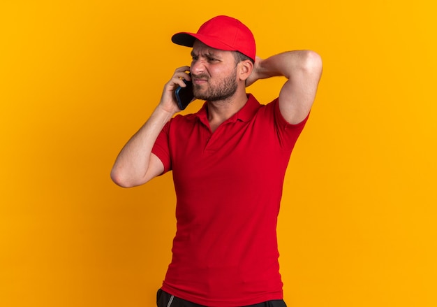 Frowning young caucasian delivery man in red uniform and cap talking on phone keeping hand behind head looking at side isolated on orange wall