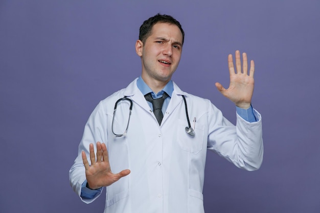 Frowning young male doctor wearing medical robe and stethoscope around neck looking at camera showing no gesture isolated on purple background