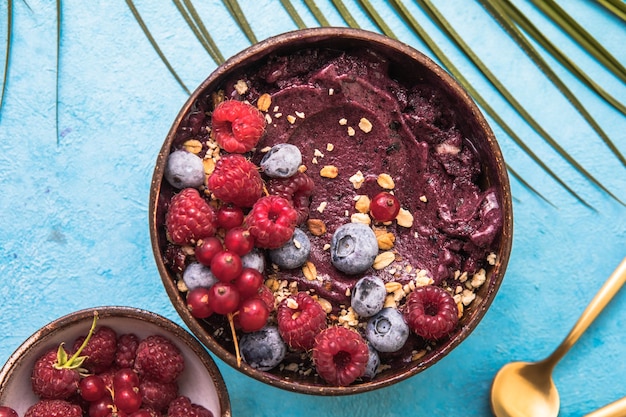 Frozen acai smoothie in coconut shell with raspberries, banana, blueberries,  fruit and granola on concrete background. Breakfast, healthy meal for summer vibes, top view, space for text