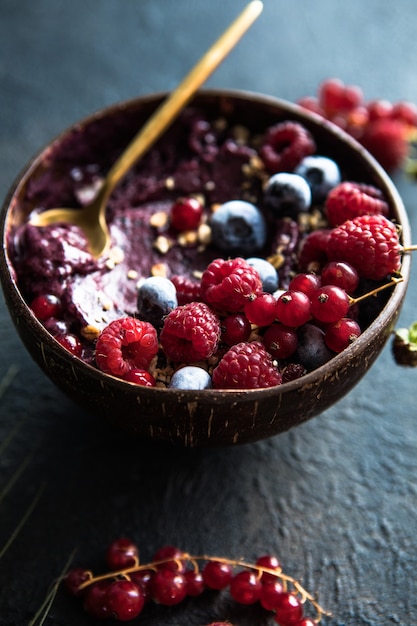 Frozen acai smoothie in coconut shell with raspberries, banana, blueberries,  fruit and granola on concrete background. Breakfast, healthy meal for summer vibes, top view, space for text