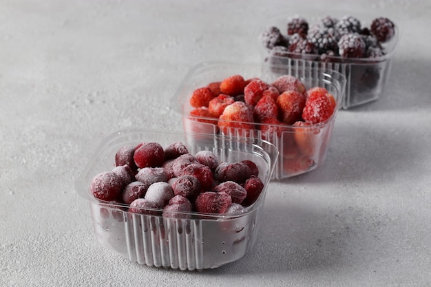 Frozen berries such as cherries, strawberries and blackberries in the storage boxes on the light gray background.