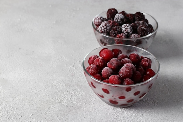 Frozen cherries and blackberries in two glass transparent bowls on light gray background. Space for text