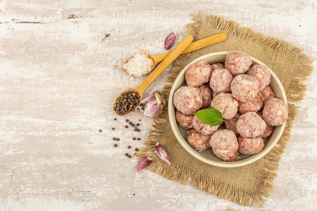 Photo frozen meatballs with spices, basil leaves, and garlic cloves in a ceramic bowl. raw ingredients for cooking food. wooden background, top view