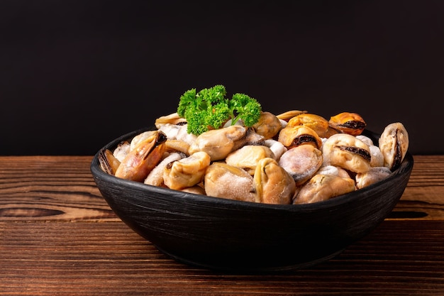 Frozen shellfish meat with green parsley in a clay black bowl on wooden table.