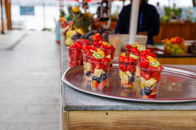 Fruit slices in transparent plastic cups with forks on a street market counter Refreshing bright fruits on a hot summer sunny day