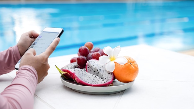 Fruit in traywith Hand of an Asian woman in a swimsuit sitting by the hotel pool Sliding mobile phone for online shopping.