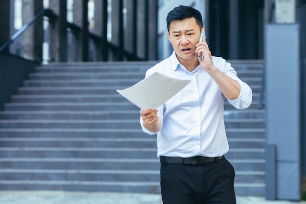 Frustrated and angry Asian businessman talking on the phone holding a letter and documents from a bank outside the office