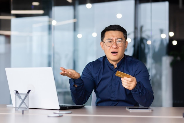 Frustrated angry asian man inside office mature boss looking at camera and holding bank credit card