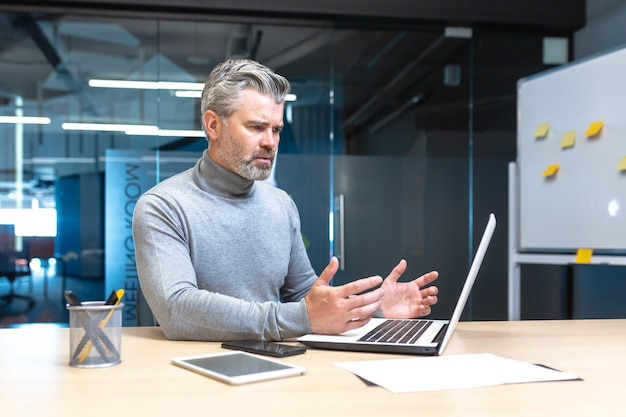 Frustrated and angry senior grayhaired businessman looking at laptop screen male manager working in modern office