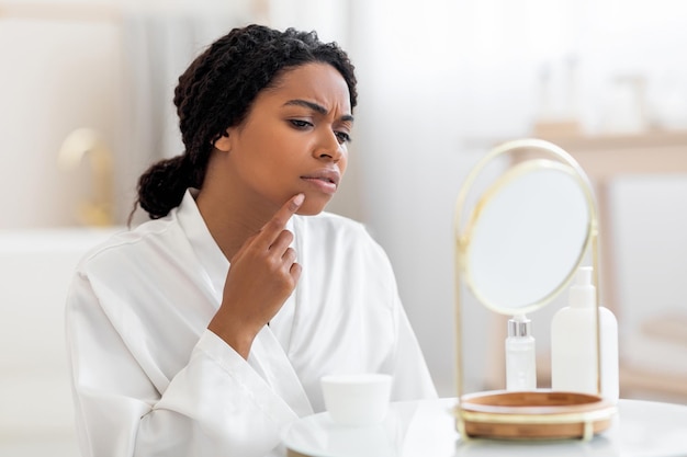 Frustrated black woman with problem skin looking at pimple on cheen