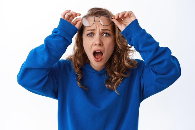 Frustrated blond girl take off glasses, gasping, looking shocked and upset at something disappointing, standing over white background