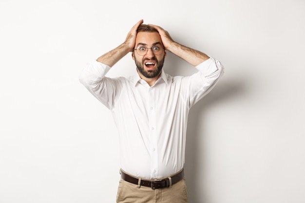 Frustrated businessman holding hands on head, looking shocked and anxious, standing over white background.